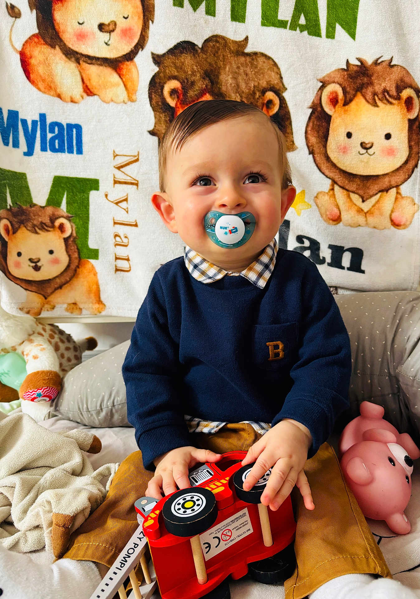 Mylan participe au concours pour gagner de l'argent avec cette photo : toddler, child, pacifier, toy, fire_truck, plush_toy, blanket, lion, smiling, indoor, cute, happy, clothing, sitting, baby, face, hands, furniture, playtime, soft_toys