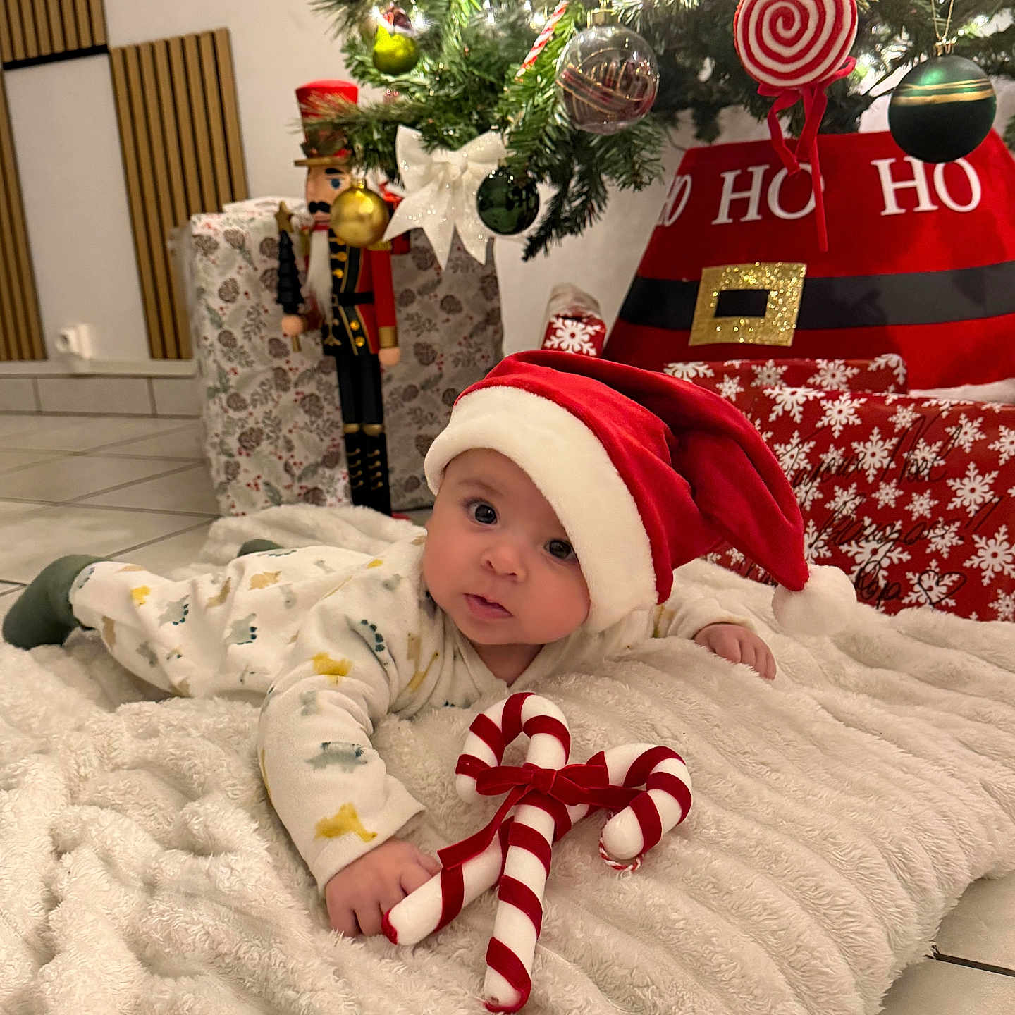 Isaï participe au concours pour gagner de l'argent avec cette photo : baby, blanket, candy_cane, christmas_tree, cute, decorations, face, festive, floor, green, holiday, indoors, infant, ornaments, presents, red, santa_hat, tiled_floor, toy, white