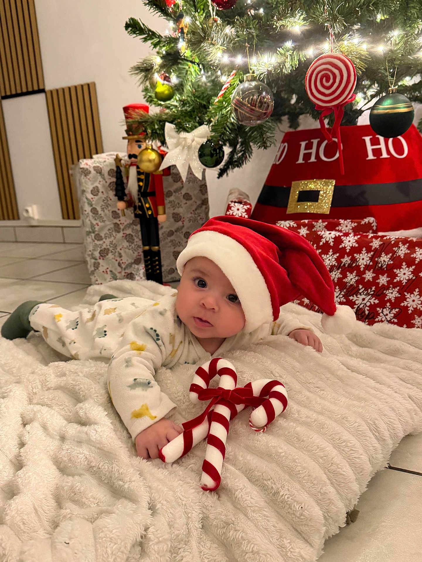 Isaï participe au concours pour gagner de l'argent avec cette photo : baby, santa_hat, candy_cane, blanket, christmas_tree, ornaments, presents, holiday, festive, floor, tiled_floor, toy, red, white, green, face, cute, infant, indoors, decorations