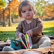 Lucy is registered to the contest to win money with this photo: child, toddler, baby, portrait, sitting, pouting, serious_expression, book, books, reading, outdoor, park, autumn, fall, leaves, blanket, sunlight, nature, brown_clothing, colorful_books