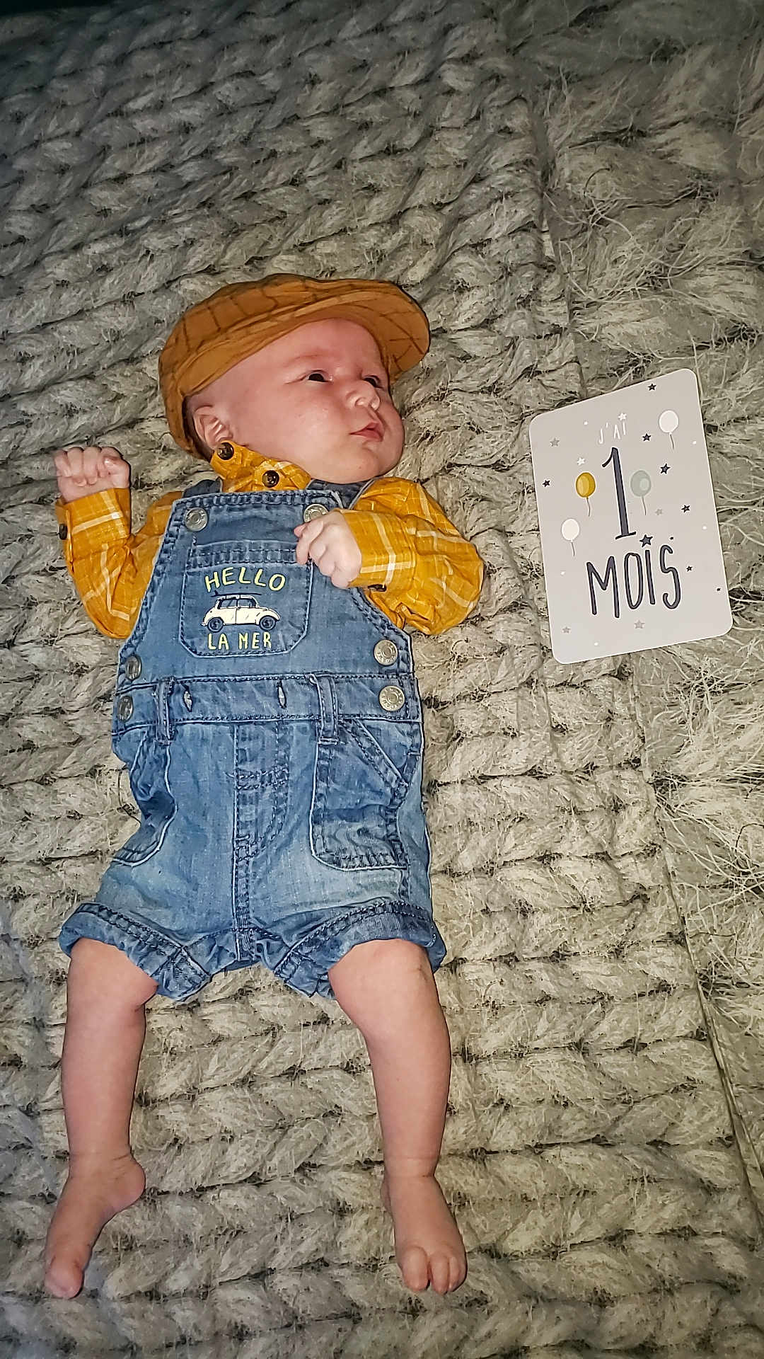 Liam participe au concours pour gagner de l'argent avec cette photo : baby, baby_clothes, blanket, cap, card, celebration, child, cute, denim_overalls, indoor, infant, lying_down, milestone, newborn, one_month, person, plaid, portrait, soft_texture, yellow_shirt