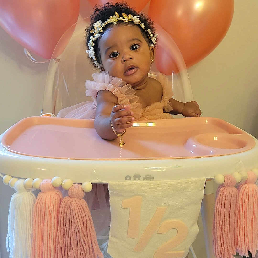 Jaior is registered to the contest to win money with this photo: baby, balloon, bracelet, celebration, child, cute, decor, dress, floor, footrest, headband, high_chair, indoor, infant, milestone, peach, person, pink, portrait, tassel