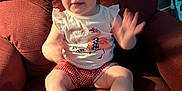 Blaze is registered to the contest to win money with this photo: baby, child, sitting, armchair, red, white, shoes, socks, shorts, tshirt, smiling, motion_blur, indoor, furniture, face, person, cute, happy, waving, plush