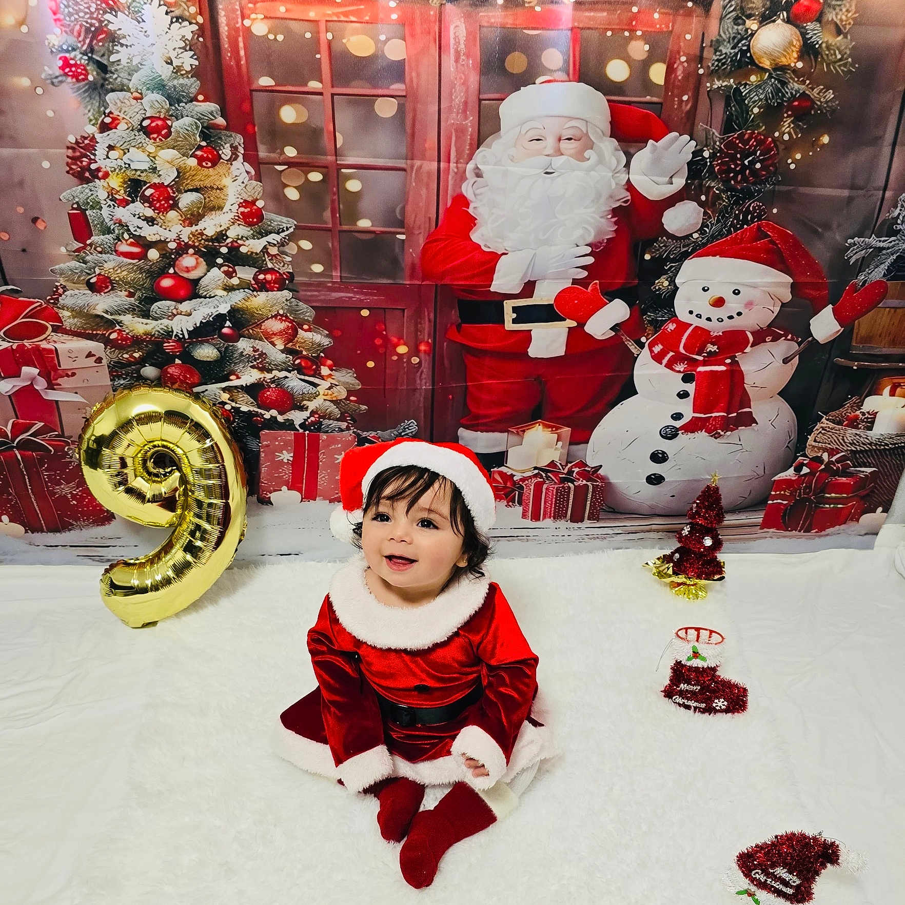Samira is registered to the contest to win money with this photo: balloon, carpet, celebration, child, christmas_outfit, christmas_theme, christmas_tree, cute, festive, gift_boxes, holiday_decor, indoor, number_9, red_clothing, santa_claus, santa_hat, smiling, snowman, toddler, white_fur_trim