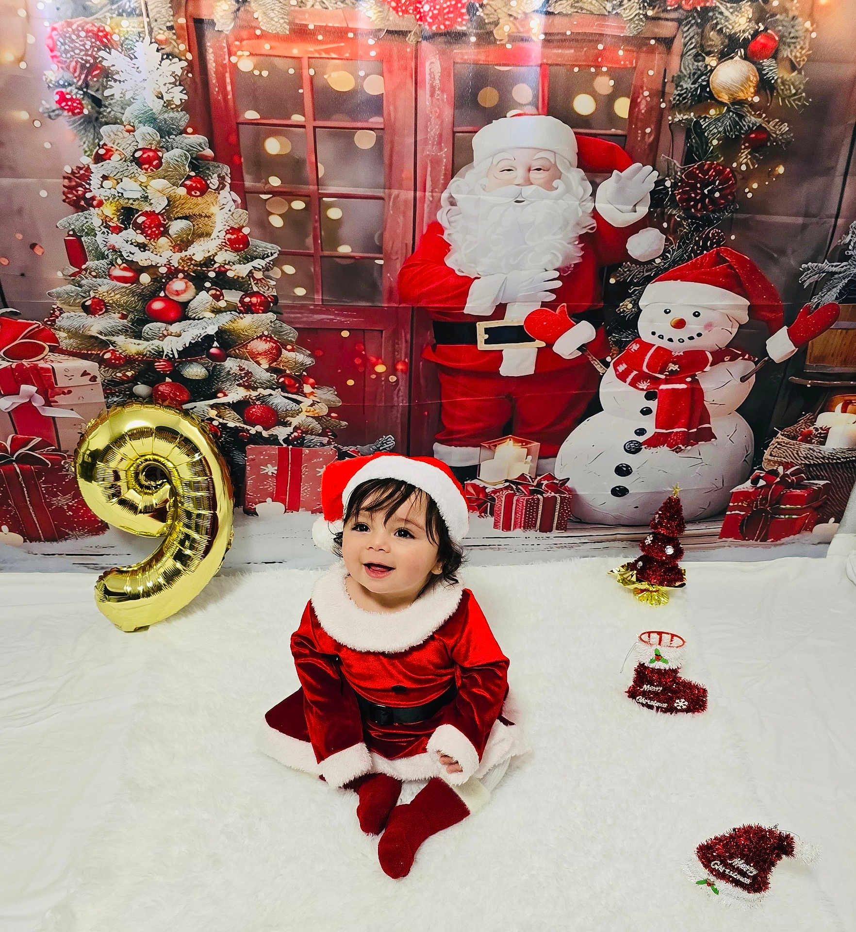 Samira is registered to the contest to win money with this photo: toddler, child, santa_hat, christmas_outfit, holiday_decor, balloon, number_9, christmas_tree, snowman, santa_claus, gift_boxes, festive, smiling, indoor, carpet, red_clothing, white_fur_trim, celebration, cute, christmas_theme