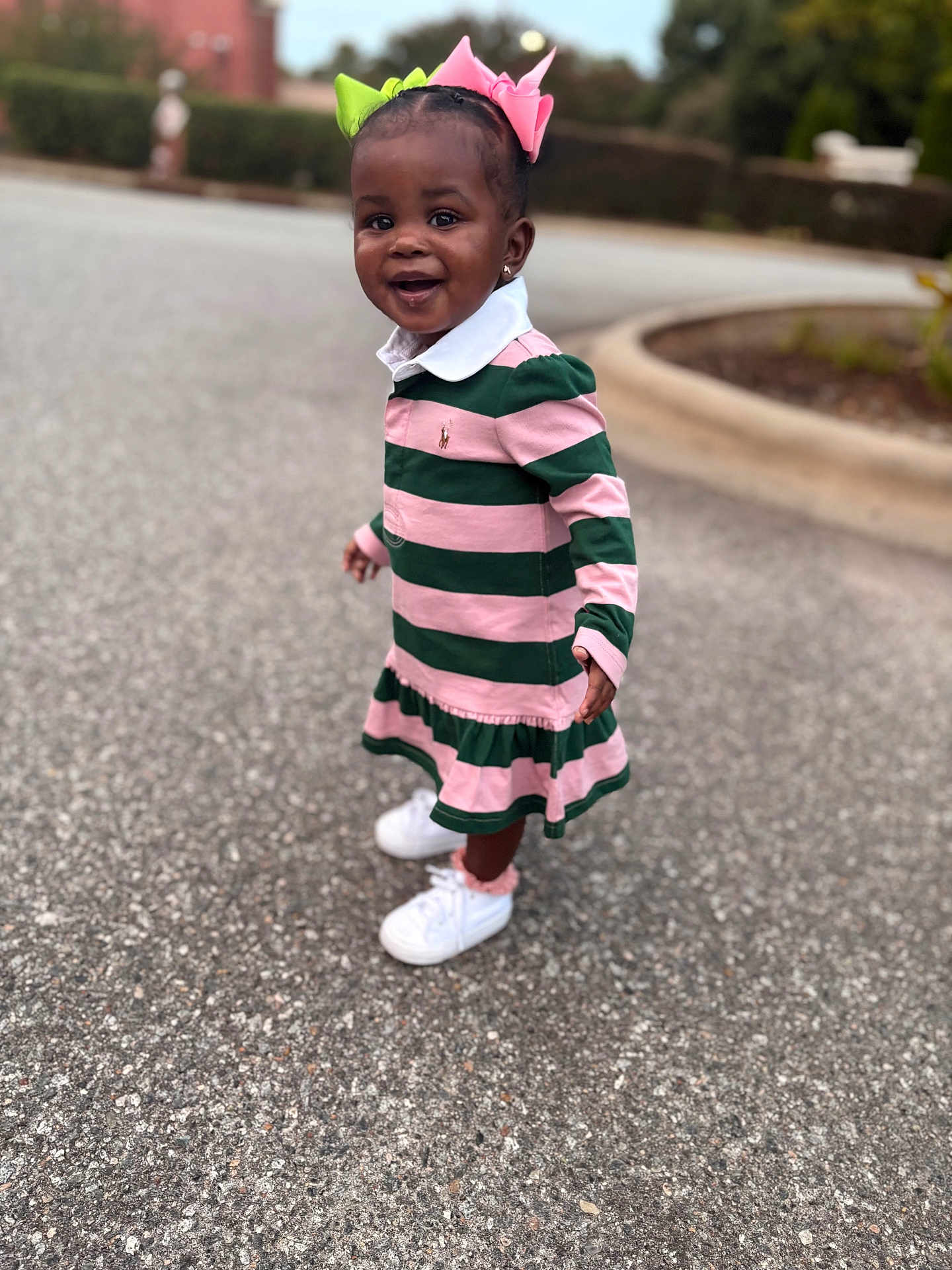 Rumani is registered to the contest to win money with this photo: toddler, child, smiling, outdoor, paved_road, dress, striped_clothing, pink, green, hair_bows, white_sneakers, earrings, happy, standing, person, curb, blurred_background, nature, daylight, fashion