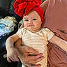 adult_hand, baby, blanket, chubby_cheeks, couch, cute, earrings, family, headband, indoor, infant, lap, living_room, onesie, person, portrait, red_bow, smile, soft_lighting, striped_clothing