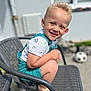 blonde_hair, casual_clothing, chair, child, crouching, curly_hair, cute, daylight, happy, metal_chair, outdoor, playful, portrait, sandals, shorts, smiling, soccer_ball, summer, sunlight, toddler