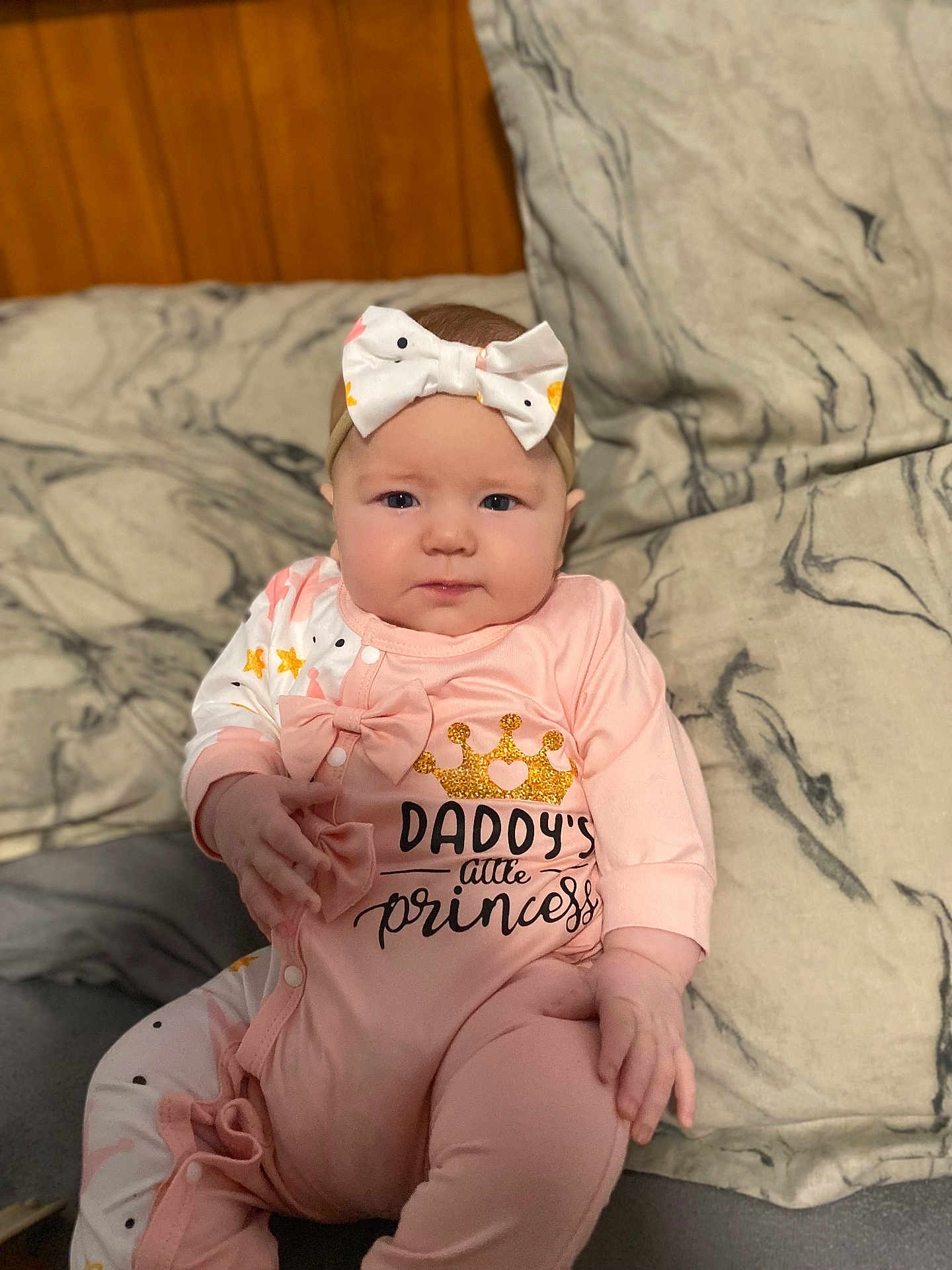Aeva is registered to the contest to win money with this photo: baby, infant, child, onesie, headband, bow, pink_clothing, crown, text_on_clothing, sitting, cushion, marble_pattern, indoor, portrait, cute, chubby_cheeks, hand, face, young_child, expression