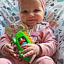 baby, bow, child, closeup, cushion, cute, face, green_toy, hand, happy, headband, indoors, infant, patterned_cushion, person, pink_clothing, playful, sitting, smiling, toy