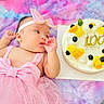 Sophia is registered to the contest to win money with this photo: baby, infant, child, birthday, cake, candles, pink_dress, hair_bow, headband, lying_down, portrait, face, hand, fruit, blueberries, orange, cream, colorful_blanket, indoor, celebration