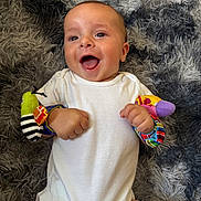 Bailey joined the competition — help win amazing prizes! baby, infant, smiling, happy, white_onesie, wrist_rattles, colorful_toys, lying_down, fluffy_rug, gray_rug, cute, child, playful, indoors, skin, face, hand, fingers, joy, expression