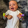baby, infant, smiling, happy, white_onesie, wrist_rattles, colorful_toys, lying_down, fluffy_rug, gray_rug, cute, child, playful, indoors, skin, face, hand, fingers, joy, expression