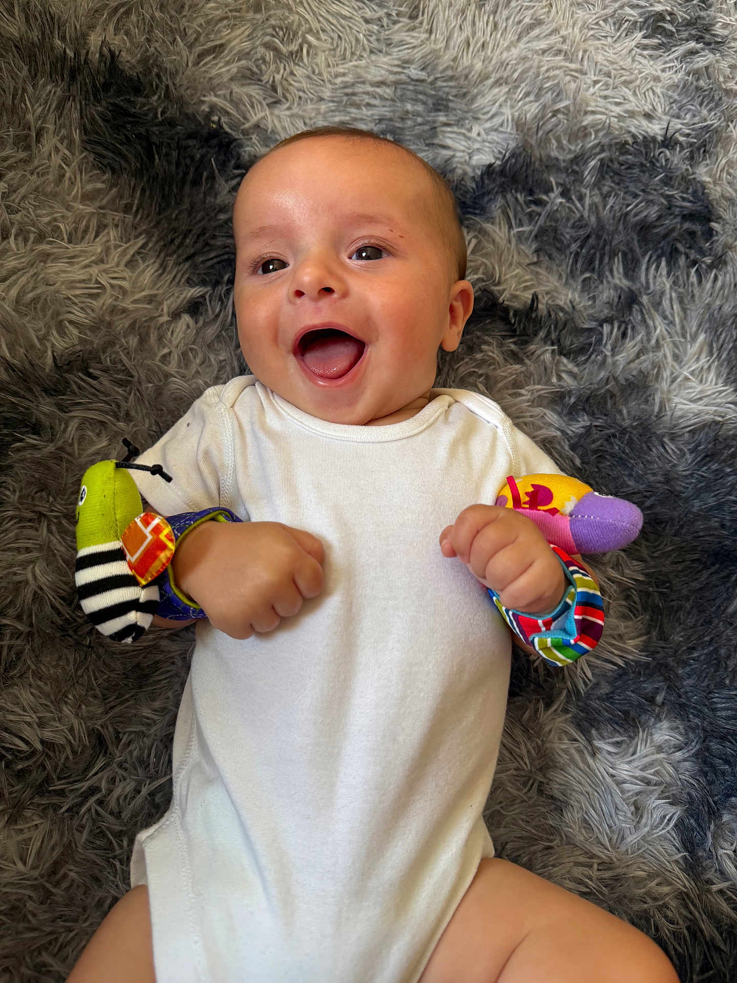 Bailey joined the competition — help win amazing prizes! baby, infant, smiling, happy, white_onesie, wrist_rattles, colorful_toys, lying_down, fluffy_rug, gray_rug, cute, child, playful, indoors, skin, face, hand, fingers, joy, expression