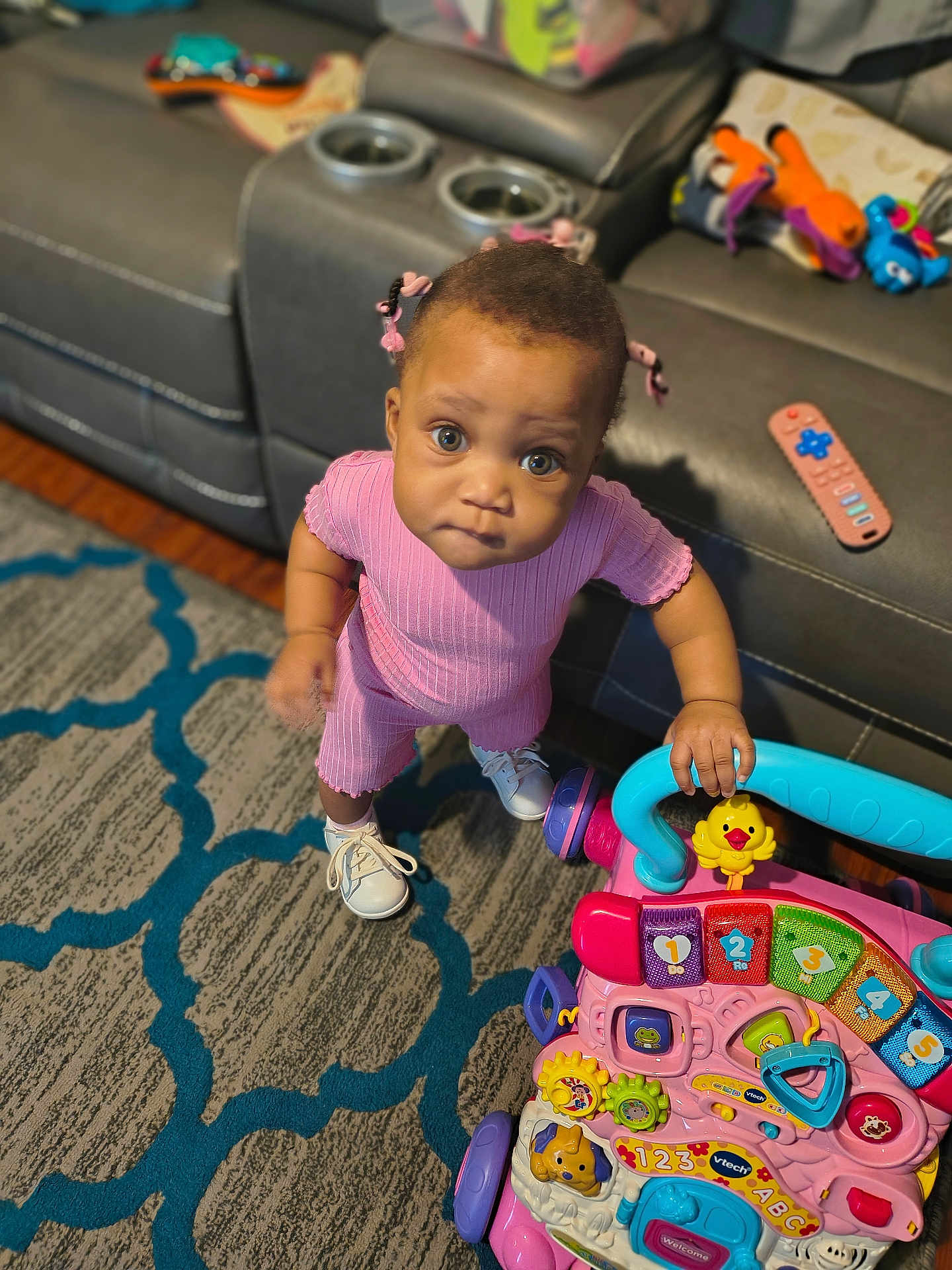 Ruby is registered to the contest to win money with this photo: toddler, child, pink_clothing, toy, walker, indoor, carpet, couch, remote_control, colorful, play, curious, standing, footwear, hair_bows, living_room, patterned_rug, plastic_toy, baby_shoes, young_child