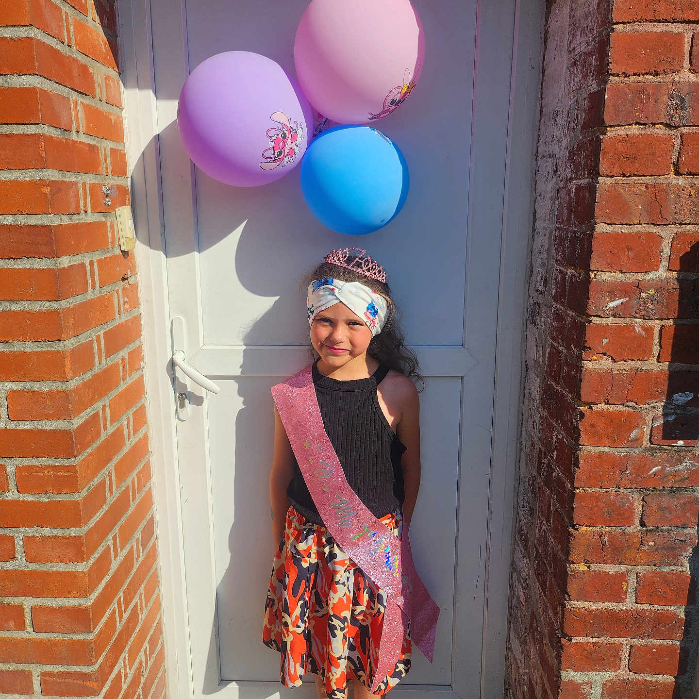 Laura a rejoint le concours — aidez-le/la à gagner de superbes lots ! balloons, brick_wall, celebration, child, clothing, colorful, door, festive, girl, happy, headband, outdoor, portrait, sash, shoes, skirt, smile, standing, sunlight, tiara