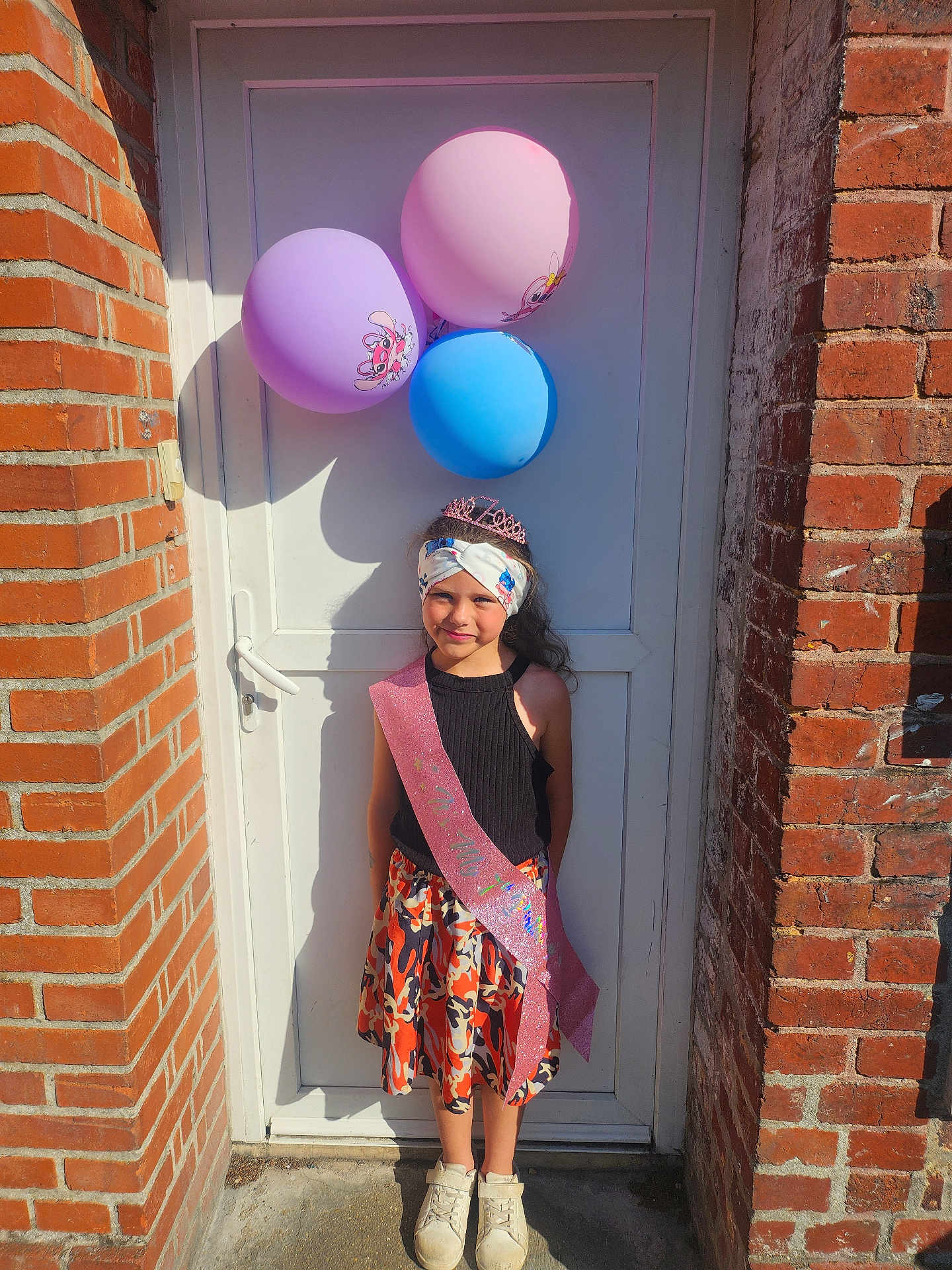 Laura a rejoint le concours — aidez-le/la à gagner de superbes lots ! child, girl, balloons, door, brick_wall, sash, headband, tiara, skirt, shoes, outdoor, sunlight, smile, celebration, festive, colorful, standing, portrait, happy, clothing