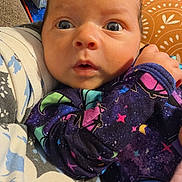 Mackenzie is registered to the contest to win money with this photo: baby, infant, child, portrait, wide_eyed, indoors, sitting, blanket, cushion, colorful_clothing, star_pattern, purple_outfit, brown_hair, blue_eyes, face, eyes, nose, mouth, home, close_up
