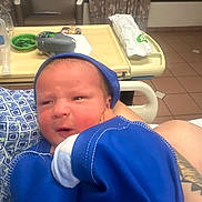 Caden is registered to the contest to win money with this photo: newborn, baby, infant, blue_clothing, hospital_room, medical_equipment, chair, blanket, person, portrait, indoors, cradled, face, headwear, human, newborn_baby, tiled_floor, curtains, arm, tattoo