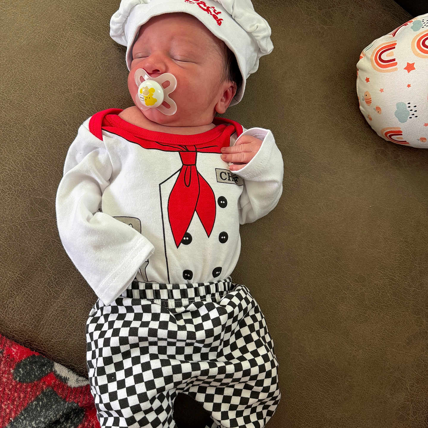 Vash is registered to the contest to win money with this photo: baby, brown_surface, checkered_pants, chef_costume, child, clothing, cushion, cute, hat, indoors, infant, newborn, pacifier, portrait, rainbow_pattern, red_scarf, resting, sleeping, soft_lighting, star_pattern