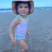 Gabriella is registered to the contest to win money with this photo: child, toddler, beach, sand, hat, swimwear, smiling, portrait, outdoor, sea, water, waves, footwear, happy, summer, curly_hair, playful, sunhat, person, vacation