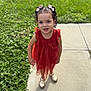 child, girl, curly_hair, pigtails, bows, orange_dress, sandals, sidewalk, grass, outdoor, sunlight, smiling, cute, standing, portrait, young, happy, nature, daytime, playful