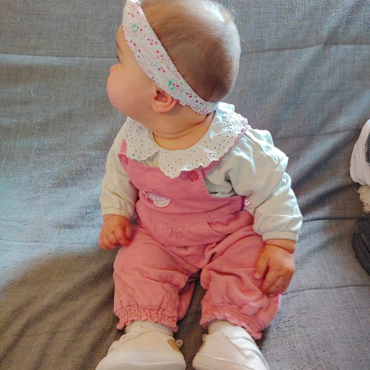 Catalina a rejoint le concours — aidez-le/la à gagner de superbes lots ! baby, child, infant, pink_overalls, headband, white_shirt, sitting, couch, gray_couch, footwear, shoes, indoors, portrait, side_view, cute, curious, small_hands, soft_lighting, casual_clothing, young_child