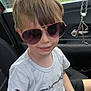 child, sunglasses, car_seat, white_tshirt, black_shorts, short_hair, smile, pen_marks, seatbelt, car_interior, window, fence, outdoor, daylight, casual_clothing, boy, portrait, happy, young, cool