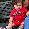 Jaziel is registered to the contest to win money with this photo: adult_arm, bench, bracelet, casual_wear, child, concrete, curly_hair, daylight, greenery, metal_bench, outdoor, person, portrait, red_clothing, serious_expression, shoes, shorts, sitting, toddler, young_child