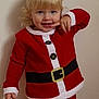 child, toddler, smiling, blonde_hair, blue_eyes, santa_outfit, red_clothing, holiday, festive, portrait, indoor, cute, happy, person, standing, long_sleeves, pants, button, belt, wall
