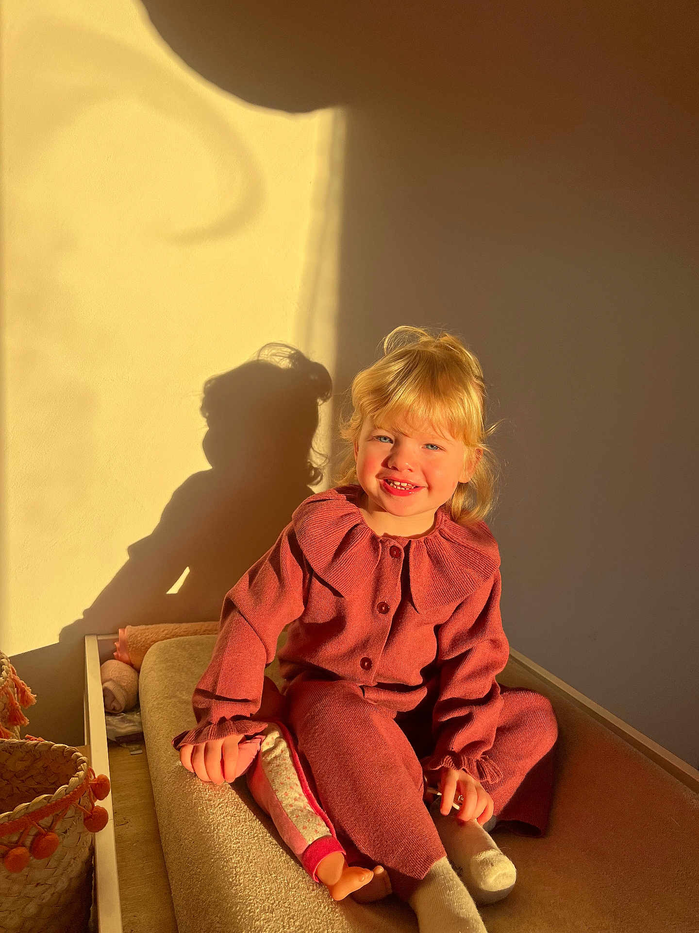 Aleya a rejoint le concours — aidez-le/la à gagner de superbes lots ! basket, blonde_hair, child, cozy, cushion, doll, happy, indoor, knitwear, person, pink_clothing, portrait, relaxed, seated, shadow, smile, sunlight, toy, wall, warm_light