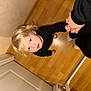 adult_hand, black_clothing, blonde_hair, blue_eyes, candid, casual, child, curious, door, flooring, holding_hands, home, indoor, looking_up, person, portrait, toddler, unposed, wooden_floor, young_child
