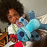 Jaciah joined the competition — help win amazing prizes! child, toddler, portrait, smiling, curly_hair, dark_skin, plush_toy, stuffed_animal, blue_plushie, stitch_character, toy, bedroom, bed, indoors, sitting, casual_clothing, red_toy, wooden_furniture, playful, photography