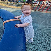 Nova is registered to the contest to win money with this photo: toddler, child, playground, outdoor, smiling, red_hair, white_dress, blue_play_structure, park, people, sunny, grass, trees, fence, casual_clothing, stroller, play_equipment, daytime, happy, person