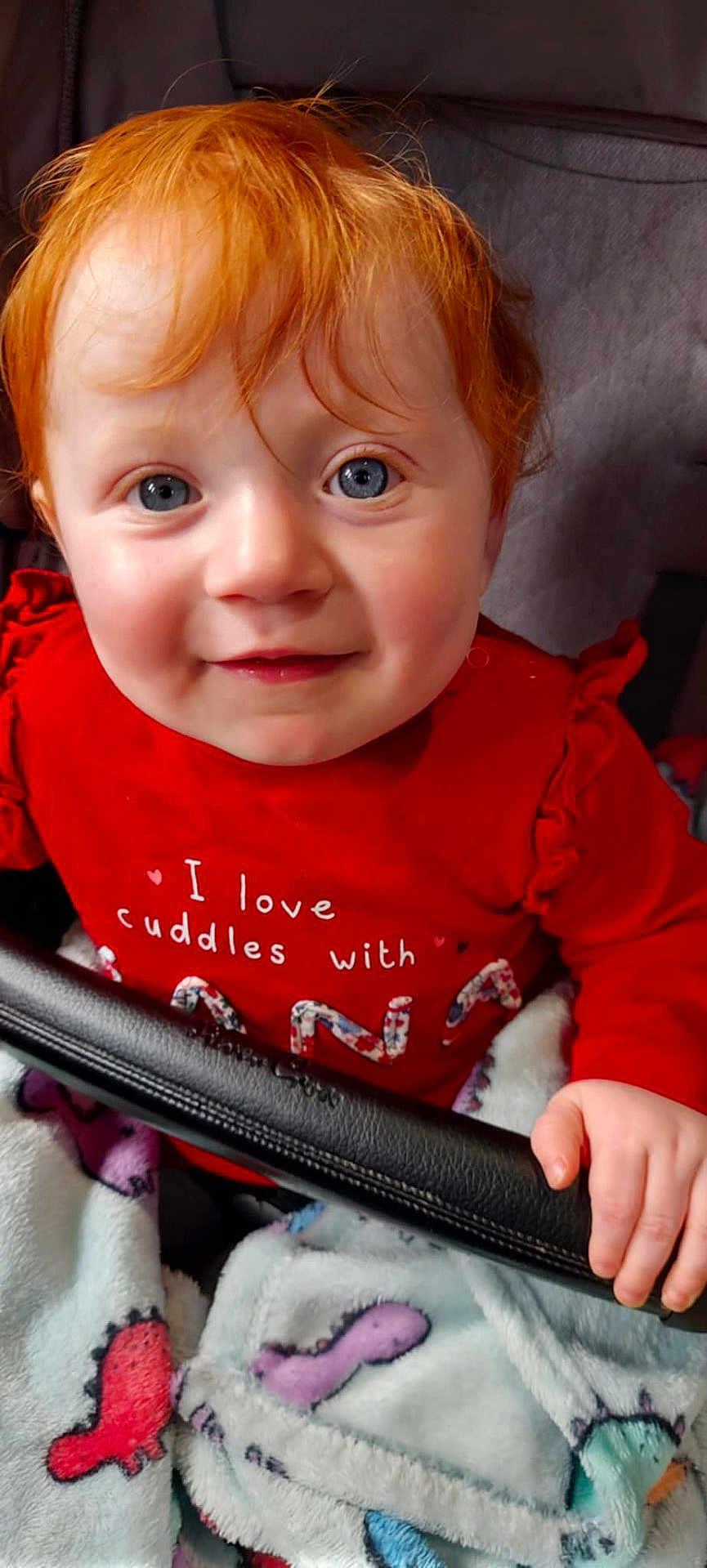 Nova is registered to the contest to win money with this photo: baby, child, red_hair, blue_eyes, smiling, face, hand, blanket, red_shirt, text_on_clothing, indoor, cute, portrait, young_child, infant, seated, holding, cozy, colorful, closeup