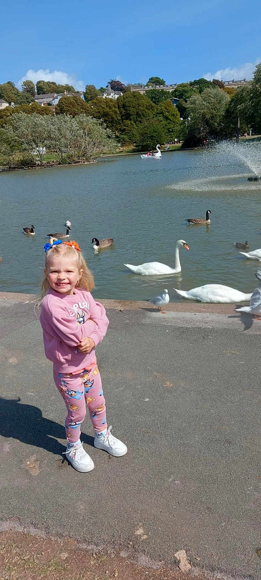 Freya is registered to the contest to win money with this photo: child, girl, smiling, outdoor, pond, water, swan, duck, birds, trees, blue_sky, clouds, grass, path, casual_clothing, shoes, sunny, nature, park, happy