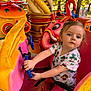 child, toddler, amusement_ride, steering_wheel, colorful, pink, yellow, outdoor, daylight, trees, playful, cartoon_animal, flamingo, elephant, seatbelt, casual_clothing, short_sleeves, sneakers, portrait, curious