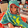 baby, blanket, child, clothing, curious, dinosaur, face, folded_clothes, furniture, home, hooded_towel, indoor, orange, person, polka_dots, seated, soft_lighting, table, towel, window