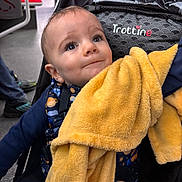 Theo participe au concours pour gagner de l'argent avec cette photo : baby, blanket, stroller, child, face, clothing, indoor, public_transport, person, yellow, blue, curious, infant, seated, expression, cute, young, transport, fabric, casual