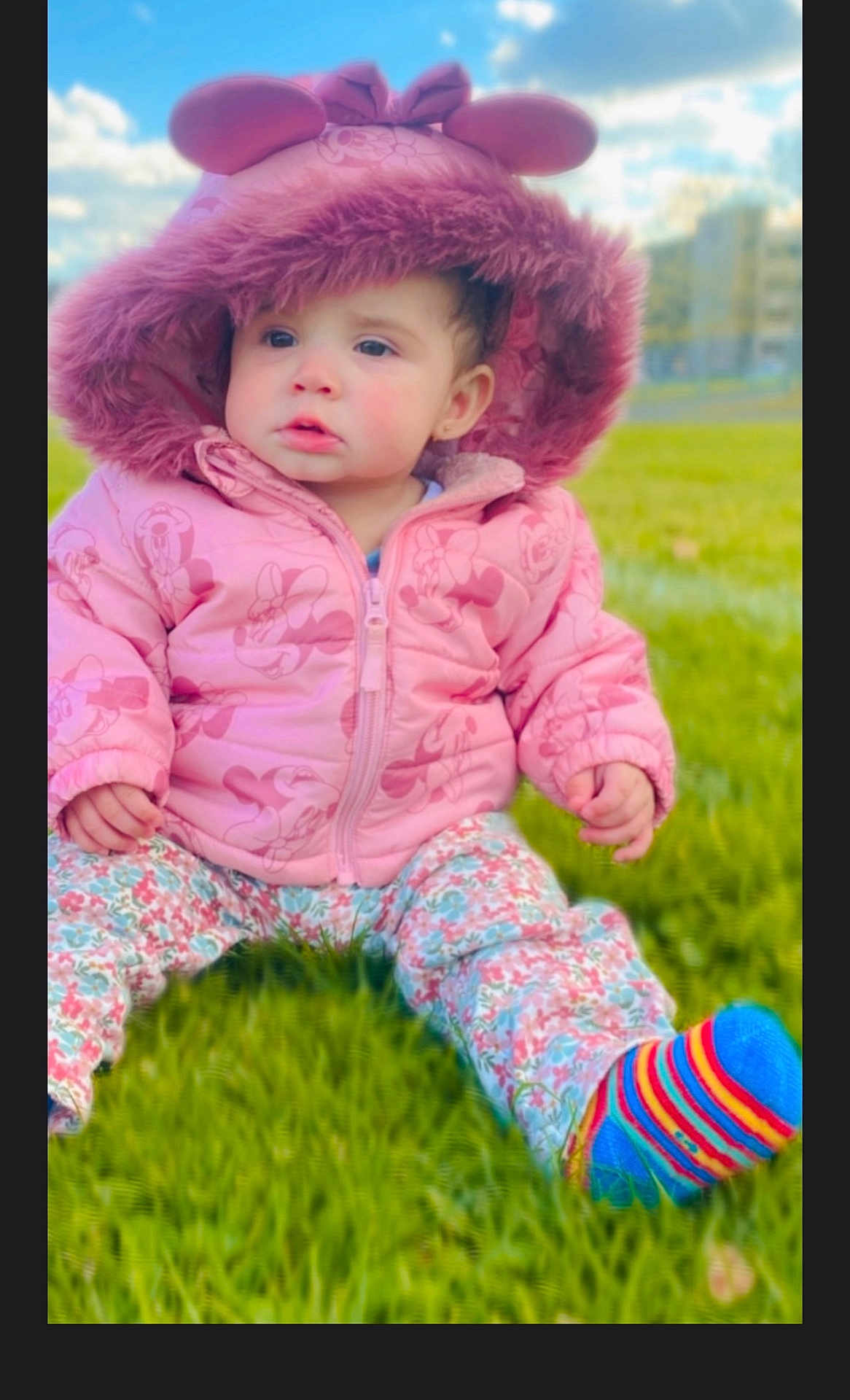 Jesenia Lemus joined the competition — help win amazing prizes! toddler, child, pink_jacket, hood, mouse_ears, grass, outdoor, clouds, sky, colorful_socks, striped_socks, floral_pants, cute, winter_clothing, baby, portrait, sitting, daylight, nature, young_child