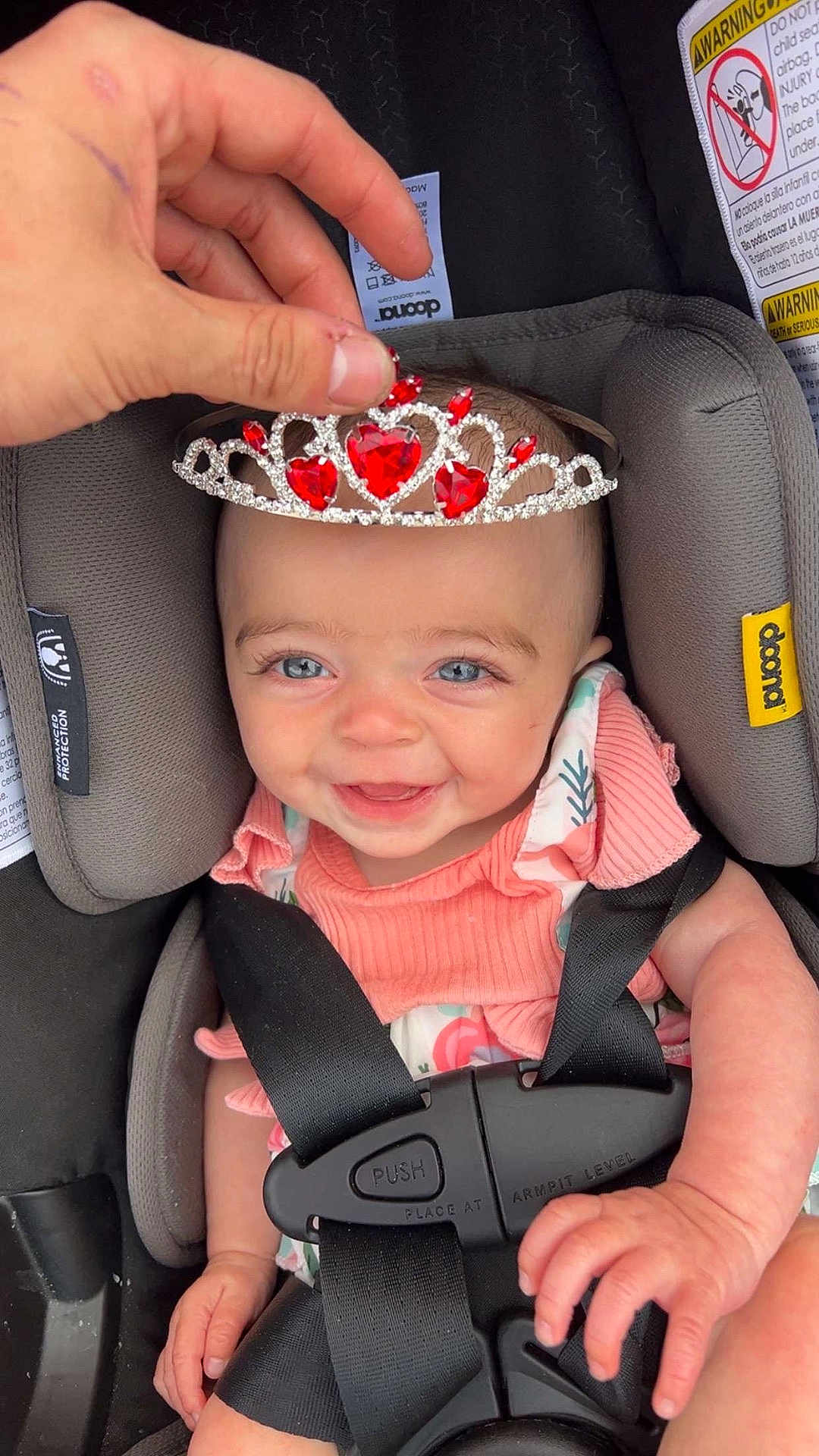 Juniper is registered to the contest to win money with this photo: baby, child, car_seat, seatbelt, tiara, hand, pink_clothing, smiling, blue_eyes, infant, safety, cute, jewelry, accessory, person, closeup, indoor, happy, portrait, seat