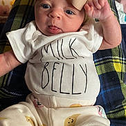 Juniper is registered to the contest to win money with this photo: baby, infant, bow, headwear, shirt, milk_belly, tongue_out, raised_fist, plaid_blanket, pants, moon_pattern, cute, playful, indoors, person, child, smiling, clothing, cozy, closeup