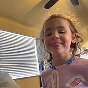 Lorelai joined the competition — help win amazing prizes! child, indoor, ceiling_fan, window, blinds, curly_hair, pink_shirt, bunny_design, tongue_out, playful, person, smiling, casual_clothing, home, light, furniture, sitting, portrait, young_child, happy