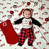 baby, blanket, child, christmas, christmas_theme, cozy, cute, festive, holiday, holiday_decor, infant, joy_text, lying_down, ornaments, plaid_pants, red_color, santa_hat, smiling, stocking, white_color