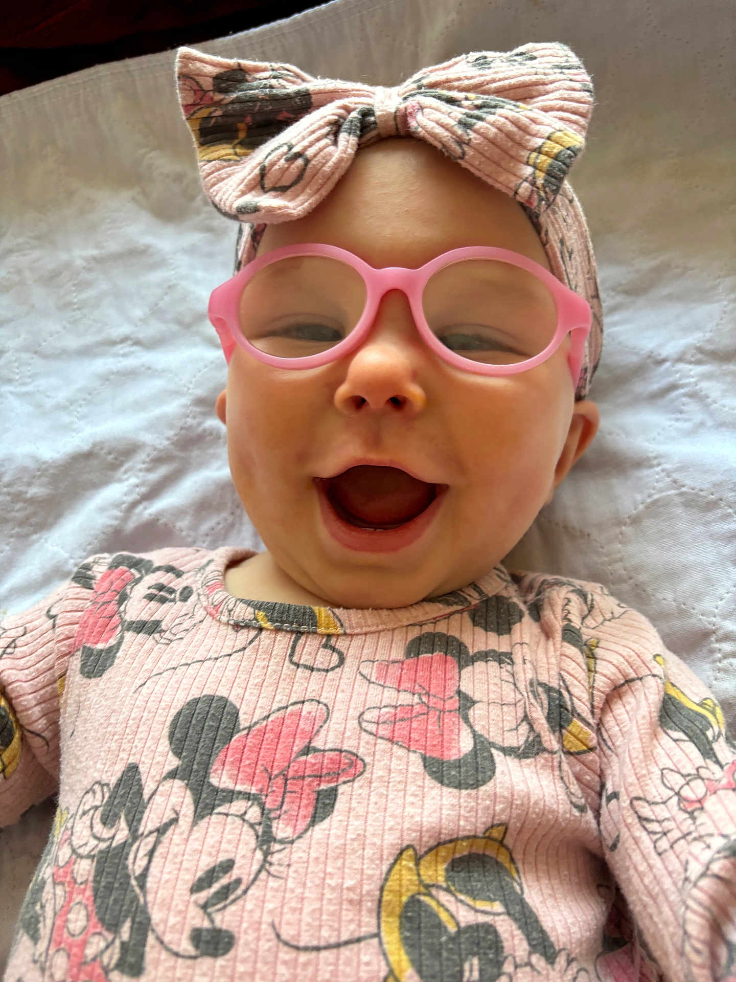 Serenity is registered to the contest to win money with this photo: baby, pink_glasses, headband, bow, minnie_mouse, clothing, laughing, happy, face, infant, cute, patterned_shirt, lying_down, white_blanket, portrait, child, smiling, apparel, accessory, closeup