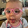 baby, child, glasses, pink_glasses, tongue_out, close_up, face, cute, indoor, living_room, tshirt, playful, person, head, expression, home, infant, young_child, casual_clothing, funny_face