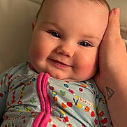 Millie is registered to the contest to win money with this photo: baby, child, face, smile, pajamas, hand, tattoo, skin, closeup, infant, cute, cheeks, person, indoors, expression, young, cozy, comfort, portrait, sleepwear