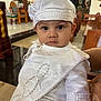 Mateo is registered to the contest to win money with this photo: baby, infant, child, christening, baptism_ceremony, white_clothing, hat, cap, lace, embroidery, church_interior, altar, pew, portrait, big_eyes, innocence, hand, wooden_furniture, ceremony, formal_outfit