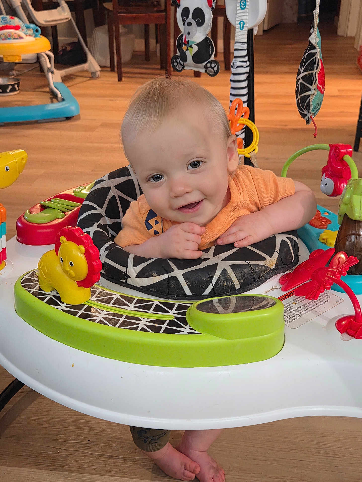 Ryker is registered to the contest to win money with this photo: baby, child, infant, activity_center, toy, yellow_lion_toy, panda_toy, orange_shirt, smiling, happy, indoor, wooden_floor, feet, playtime, toddler, colorful, seat, baby_toys, cute, face