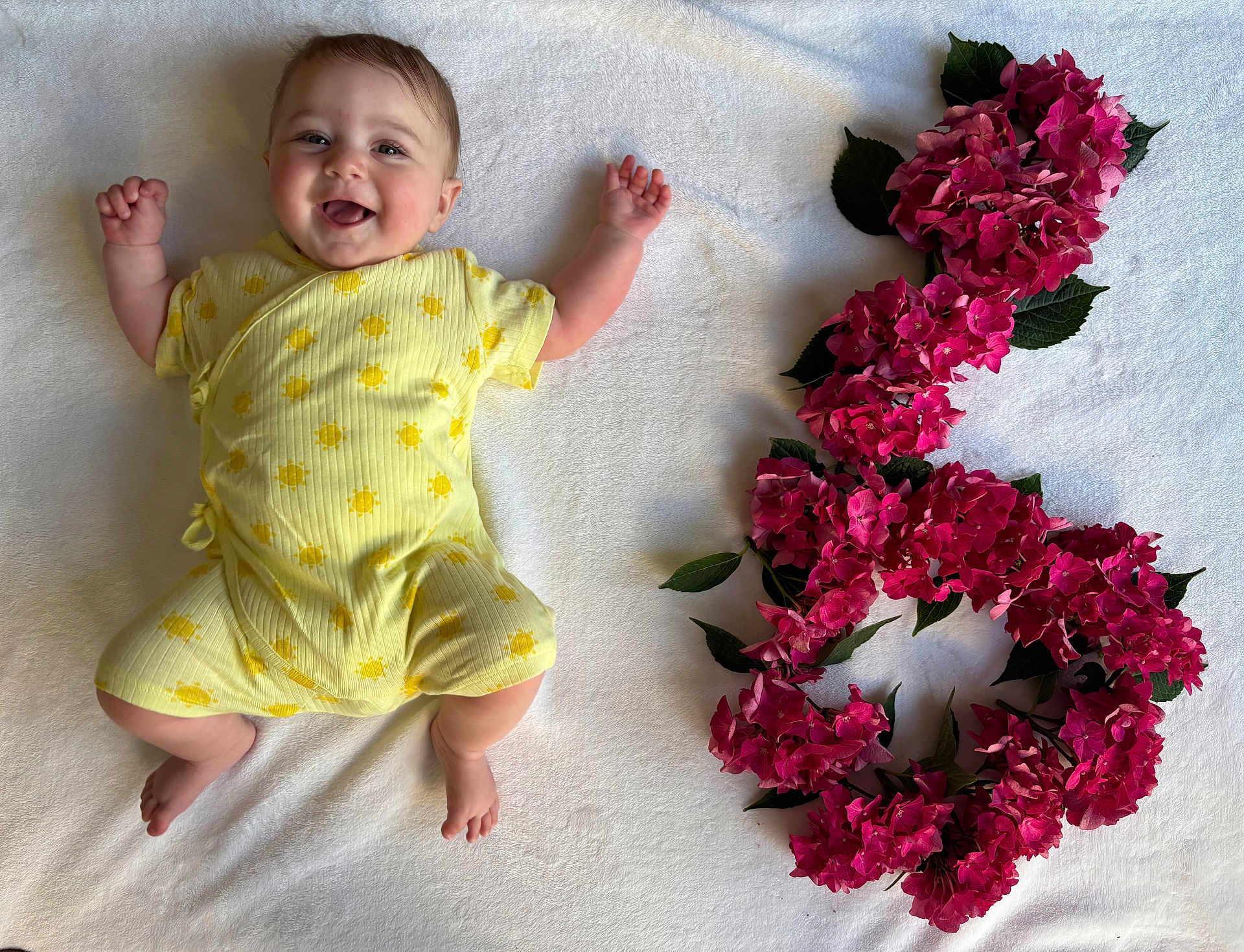 Lola a rejoint le concours — aidez-le/la à gagner de superbes lots ! baby, child, smiling, yellow_outfit, sun_pattern, pink_flowers, number_six, white_blanket, happy, portrait, infant, cute, celebration, floral_arrangement, laying_down, arms_up, small_feet, skin, person, indoors